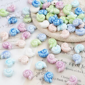 May include: A selection of ceramic seashell beads in pastel shades of blue, pink, green, and purple. The beads are scattered on a white surface, with some on a woven basket. The beads feature a spiral design.
