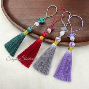 May include: Four decorative tassels in teal, red, gray, and lavender. Each tassel features a matching cord loop, a decorative knot, a white bead, and a gold-colored band securing the silk-like threads. The tassels are arranged in a row.
