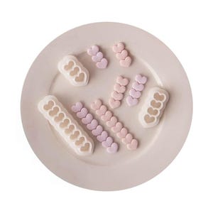 Puede incluir: Un conjunto de moldes de silicona y formas moldeadas con diseño de corazones conectados. El set incluye dos moldes blancos con cavidades en forma de corazón y varias filas de pequeñas formas de corazón en tonos pastel rosa, lavanda y melocotón.