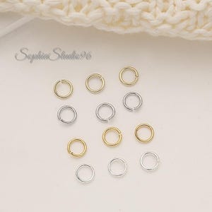 May include: A collection of gold and silver-colored jump rings, scattered on a light-colored surface. The rings are small and circular, with a split for attaching to other jewelry components. The image includes the text "Sophia Studio96".
