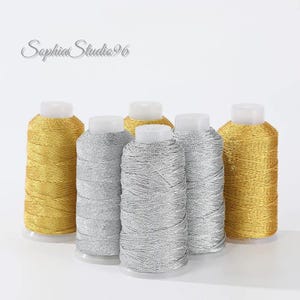 Peut inclure: Cinq bobines de fil à broder métallique disposées en groupe, comprenant deux bobines dorées aux extrémités et trois bobines argentées au centre. Chaque bobine possède un noyau en plastique blanc. Le texte SophiaStudio96 est visible en haut à gauche.