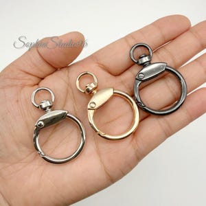 May include: Three circular metal swivel clasps in silver, gold, and gunmetal finishes. Each clasp features a round ring attached to a spring-loaded gate mechanism that opens and closes, allowing for secure attachment to various items or accessories.
