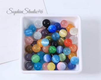 May include: A square white tray filled with a collection of round, smooth cat's eye beads in various colors including blue, green, yellow, orange, pink, black, and white. The beads feature a distinct chatoyant band across their centers.