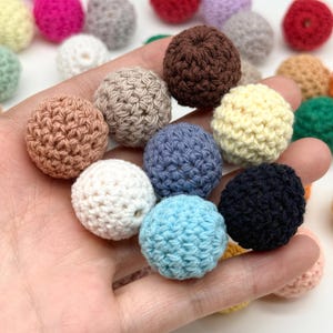 May include: A collection of small, spherical crochet beads in various solid colors including brown, beige, white, navy blue, light blue, and grey. Each bead features a textured, knotted yarn surface with a visible center hole for threading.