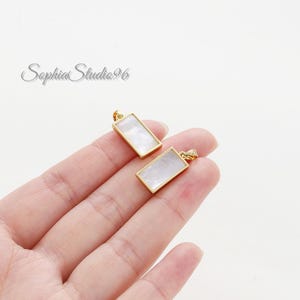 Puede incluir: Dos colgantes rectangulares con una incrustación de nácar blanco iridiscente y liso, engastados en un marco de metal dorado pulido. Cada colgante incluye una pequeña anilla integrada en la parte superior para sujetarlo a una cadena.