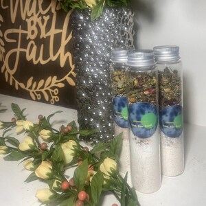 May include: Three clear glass tubes filled with white granules and dried herbs, labeled "Tried, True, True'd". A silver vase with yellow flowers and greenery is in the background, along with a wooden sign that says "Walk By Faith".