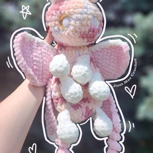 May include: A plush moth toy made of variegated pink, white, and peach yarn with large iridescent eyes. The toy features white pom-pom-like feet and hands, and wide wings. Text on the image reads Moth Snuggler and Just Mae Crochet with stars.