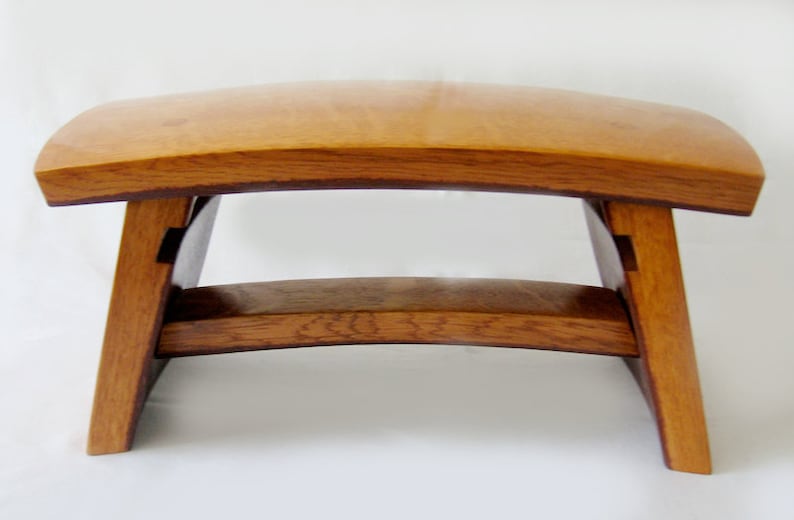 May include: A wooden stool with a curved top and a simple, minimalist design. The stool is made of light brown wood with a natural finish.