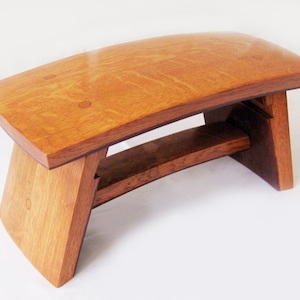 May include: A wooden bench with a curved top and a unique, angled base. The bench is made of light brown wood with a natural finish.