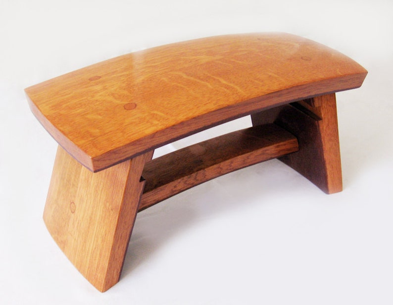 May include: A wooden bench with a curved top and a unique, angled base. The bench is made of light brown wood with a natural finish.