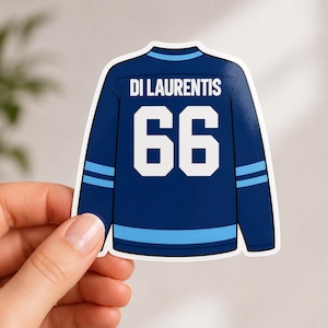 May include: A sticker of a navy blue hockey jersey with the name "DI LAURENTIS" above the number "66" in white. The jersey has light blue stripes on the sleeves and bottom. The sticker is held by a hand.