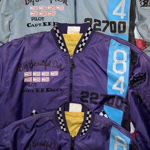 May include: Three bomber jackets in the image. One is light blue, and two are purple. Each jacket has a zipper, the text "Big Beautiful Doll", and the number "84". The light blue jacket has the number "22700".