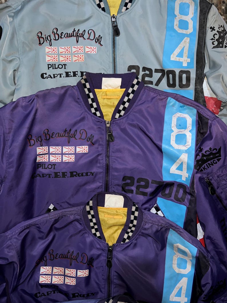 May include: Three bomber jackets in the image. One is light blue, and two are purple. Each jacket has a zipper, the text "Big Beautiful Doll", and the number "84". The light blue jacket has the number "22700".