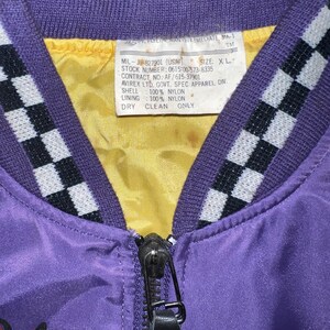 May include: Close-up of a vintage purple nylon bomber jacket with a black zipper and a yellow lining. The jacket has a checkered pattern on the collar and a tag with text. The text on the tag includes "MIL-J-827901 (USAF)" and "DRY CLEAN ONLY."