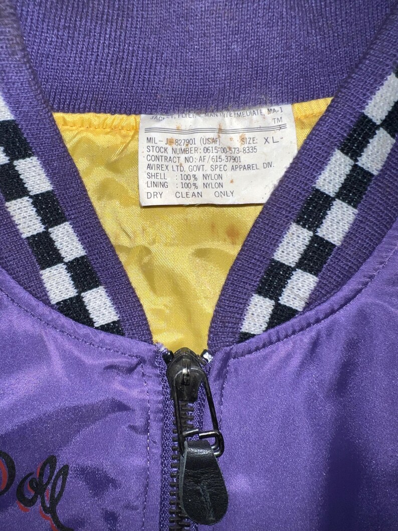 May include: Close-up of a vintage purple nylon bomber jacket with a black zipper and a yellow lining. The jacket has a checkered pattern on the collar and a tag with text. The text on the tag includes "MIL-J-827901 (USAF)" and "DRY CLEAN ONLY."