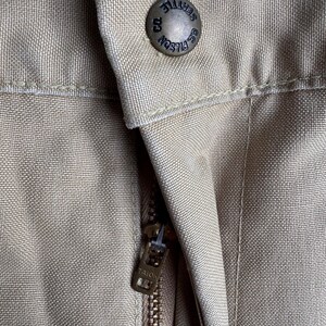 Puede incluir: Primer plano de un pantal&oacute;n color beige con un bot&oacute;n y cremallera met&aacute;licos. El bot&oacute;n est&aacute; grabado con "FILSON SEATTLE CO.". El tirador de la cremallera est&aacute; marcado "TALON". La tela tiene una apariencia texturizada.