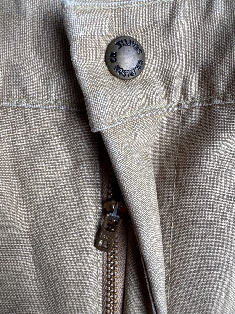 Puede incluir: Primer plano de un pantal&oacute;n color beige con un bot&oacute;n y cremallera met&aacute;licos. El bot&oacute;n est&aacute; grabado con "FILSON SEATTLE CO.". El tirador de la cremallera est&aacute; marcado "TALON". La tela tiene una apariencia texturizada.