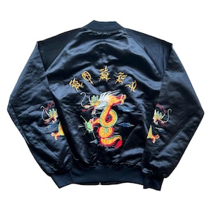 Può includere: Giacca bomber in raso nero con ricamo a forma di drago nei colori rosso, giallo e verde. La giacca presenta una chiusura frontale con zip, polsini a coste e una classica silhouette da bomber. La parte posteriore della giacca presenta un grande disegno di drago con caratteri giapponesi.