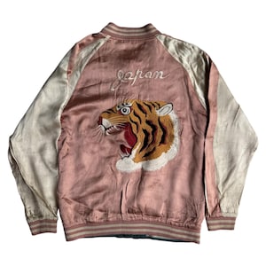 Może przedstawiać: Różowa satynowa kurtka bomberka z kremowymi rękawami. Z tyłu haftowany tygrys i napis "Japan". Kurtka ma prążkowany kołnierz i mankiety.
