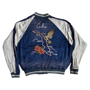 Może przedstawiać: Niebieska kurtka bomberka z białymi rękawami. Z tyłu znajduje się haftowana mapa Kuby, napis "Cuba" i ptak. Mapa zawiera miasta Hawana, Trynidad, Holguín i Santiago de Cuba. Kurtka ma zielono-biały kołnierz i mankiety w paski.