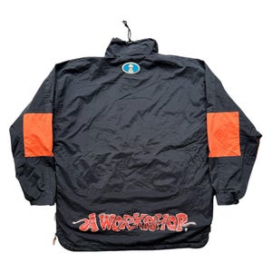 May include: Black nylon jacket with orange shoulder patches and a small alien logo. The jacket features the text "A WORKSHOP" in red script across the bottom. The jacket has a drawstring hood and a zipper closure.