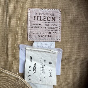 Puede incluir: Primer plano de una tela beige con dos etiquetas. La etiqueta superior dice "A Genuine Filson" con el texto "Might as well have the best" y "C.C. Filson Co. Seattle." La etiqueta inferior dice "Made in U.S.A."