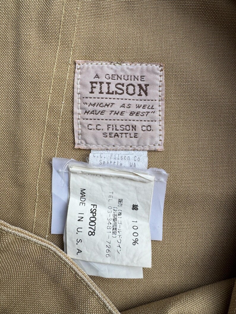 Puede incluir: Primer plano de una tela beige con dos etiquetas. La etiqueta superior dice "A Genuine Filson" con el texto "Might as well have the best" y "C.C. Filson Co. Seattle." La etiqueta inferior dice "Made in U.S.A."
