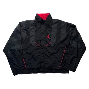 May include: Black windbreaker jacket with red detailing and a red lining. The jacket has a high collar, a front zip, and the Jumpman logo in red on the chest. The sleeves feature a striped design.