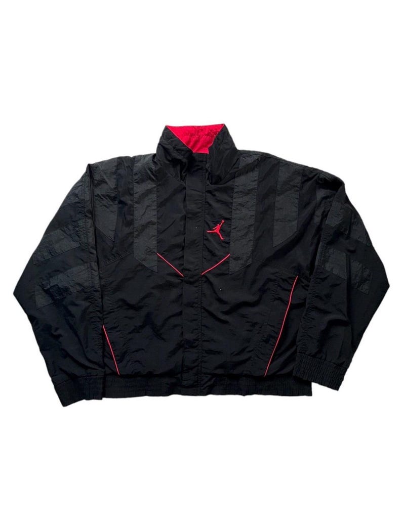 May include: Black windbreaker jacket with red detailing and a red lining. The jacket has a high collar, a front zip, and the Jumpman logo in red on the chest. The sleeves feature a striped design.