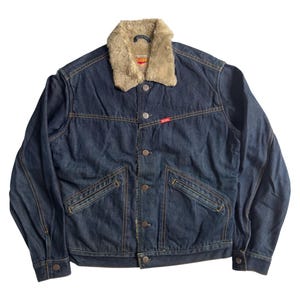May include: A dark blue denim jacket with a tan faux fur collar. The jacket features a button-up front, two chest pockets, and two side pockets. The stitching is a contrasting tan colour. A small red tag is visible on the front.