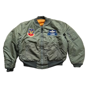 Op de afbeelding: Een groene bomberjack met een ritssluiting. De jas heeft twee patches aan de linkerkant, een met een zwaard en vleugels en de andere met het woord "LEWIS".