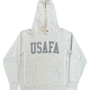 Może przedstawiać: Jasnoszara bluza z kapturem z napisem "USAFA" w ciemnoszarych blokowych literach na piersi. Bluza ma przednią kieszeń i kaptur ze sznurkiem. Tkanina wydaje się być miękkim, wygodnym materiałem.
