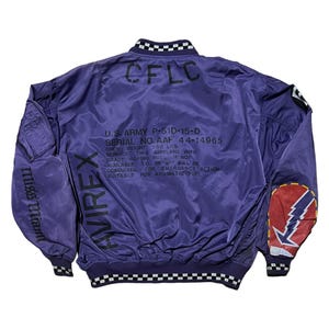 May include: Purple bomber jacket with black and white checkered trim on the collar and cuffs. The back of the jacket features text including "CFLC", "U.S. ARMY P-51D-15-D", and "AVIREX". The left sleeve has "Miss Miant" printed on it, and the right sleeve has a patch with a lightning bolt.