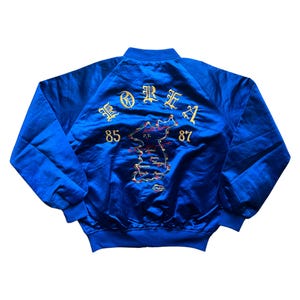 Può includere: Giacca bomber in raso blu reale con ricami dorati. Il retro presenta un disegno a mappa con la scritta "KOREA" e i numeri "85" e "87". La giacca ha colletto, polsini e orlo a coste.