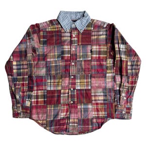 Op de afbeelding: Een button-down shirt met een patchwork design in verschillende kleuren, waaronder rood, blauw en geel. De kraag is lichtblauw en wit gestreept. Een klein zwart logo is zichtbaar op de borst.