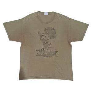 Op de afbeelding: Een beige T-shirt met een zwart-witte afbeelding van een cartoonfiguur dat in een microfoon zingt. De tekst "JEMINI" en "THE EXPERIENCE" staat onder de afbeelding.