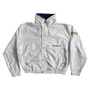 Könnte beinhalten: Weißer Vintage-Jacke mit hohem Kragen und Druckknöpfen. Sie hat ein marineblaues Futter am Kragen und die Aufschrift "LE MANS FRANCE" und "24 HOURS" auf den Ärmeln. Die Jacke hat zwei Vordertaschen und einen gerippten Saum.