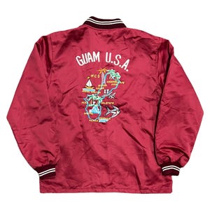 Może przedstawiać: Bordowa satynowa kurtka z biało-bordowymi paskami na kołnierzu i mankietach. Tył zdobi haftowany wzór z napisem "GUAM U.S.A." oraz kolorowa ilustracja mapy z tekstem.