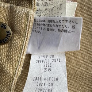 Puede incluir: Primer plano de una prenda de color canela con un bot&oacute;n met&aacute;lico que dice "CO SEATTLE". Las etiquetas adjuntas muestran texto que incluye "STYLE 78 SIZE 36" y "100% algod&oacute;n Care on reverse". La tela de la prenda tiene una apariencia texturizada.