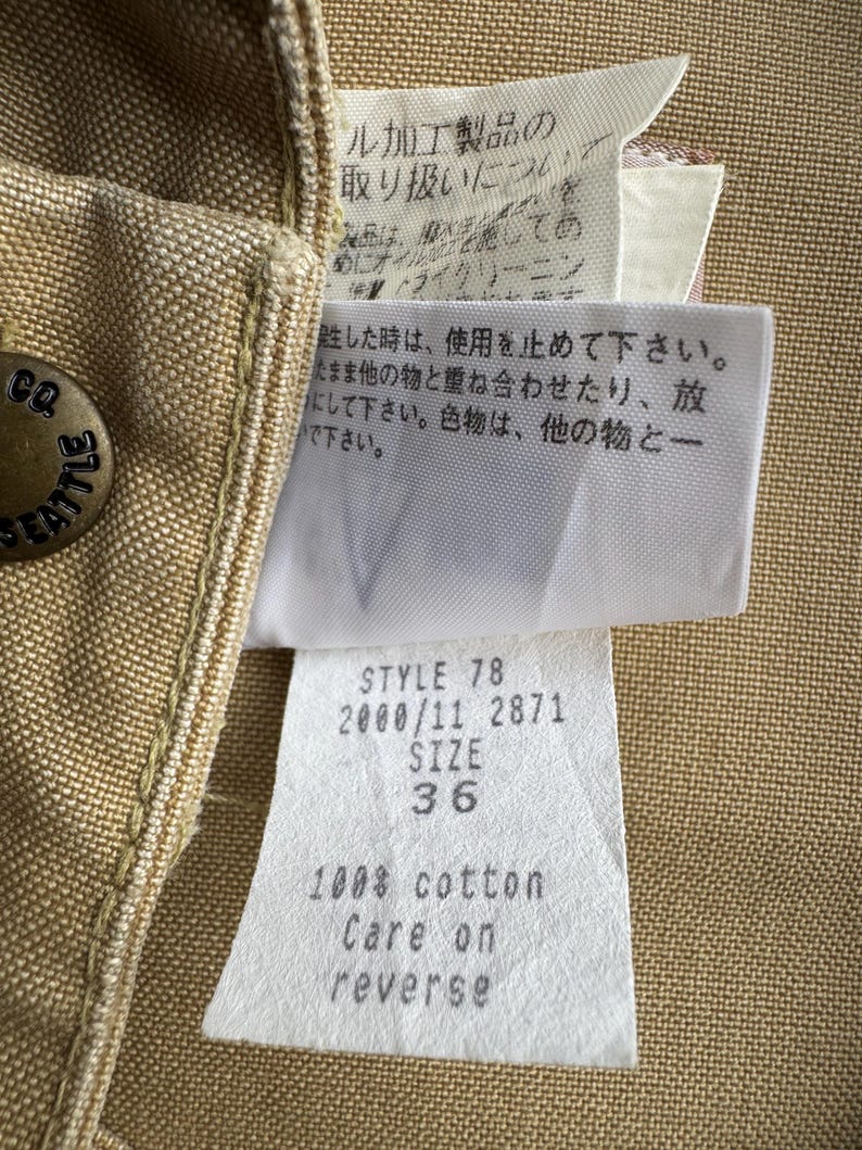 Puede incluir: Primer plano de una prenda de color canela con un bot&oacute;n met&aacute;lico que dice "CO SEATTLE". Las etiquetas adjuntas muestran texto que incluye "STYLE 78 SIZE 36" y "100% algod&oacute;n Care on reverse". La tela de la prenda tiene una apariencia texturizada.