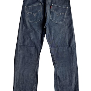 Puede incluir: Vaqueros de mezclilla azul oscuro con el diseño clásico de bolsillo trasero de Levi's. Los vaqueros tienen un corte de pierna recta y una etiqueta roja de Levi's. Las costuras son visibles en blanco. Los vaqueros se muestran sobre un fondo blanco.