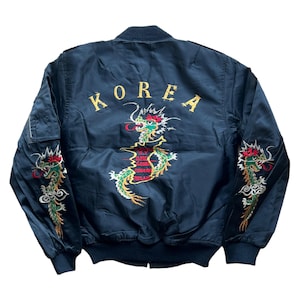 May include: Navy blue bomber jacket with embroidered dragon and "KOREA" text. The dragon is green, red, and gold. The jacket has a ribbed collar, cuffs, and hem. A pocket is visible on the left sleeve.