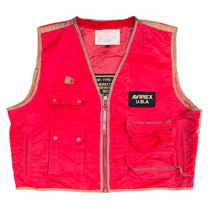 May include: Bright red vest with multiple pockets and a zipper closure. Features a gold trim and the "AVIREX U.S.A." logo. Includes a small patch with text and a label near the collar. A vintage-style garment.
