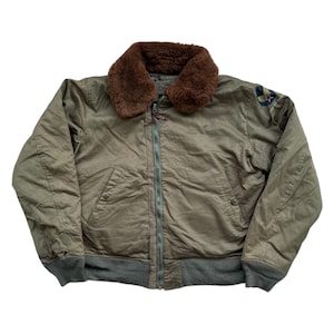 Può includere: Giacca bomber verde oliva con colletto in pelliccia marrone. La giacca presenta una chiusura con cerniera, polsini a coste e una toppa sulla manica superiore sinistra. La giacca ha un aspetto vintage e usurato.