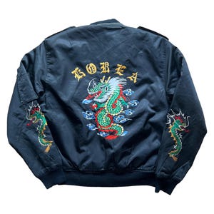 Może przedstawiać: Czarna kurtka bomberka z haftowanym smokiem i napisem "KOREA" na plecach. Smok jest zielony, czerwony i żółty z niebieskimi akcentami. Dodatkowe hafty smoków na rękawach.