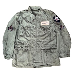 Può includere: Giacca militare verde oliva con più tasche, chiusura con bottoni e colletto. La giacca presenta toppe sulle maniche e sul petto, tra cui un'emblema a stella e il nome "OSBORN". La giacca ha un aspetto usurato.
