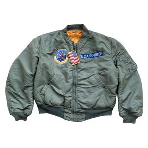 Op de afbeelding: Een groene bomberjack met een ritssluiting en twee zakken. De jas heeft een U.S. Air Force patch en een Amerikaanse vlag patch aan de linkerkant van de borst.