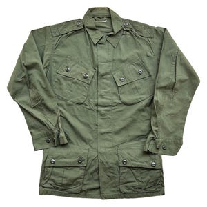 May include: An olive green military-style jacket with multiple pockets and button fastenings. The jacket has a collared neckline, long sleeves, and epaulettes on the shoulders. The fabric looks to be a lightweight cotton or blend, suitable for warmer weather.