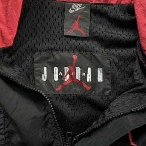 May include: Black and red Nike Jordan jacket. The jacket has a black mesh lining, a red collar, and a zip closure. The Nike logo and the Jordan logo are visible on the jacket.