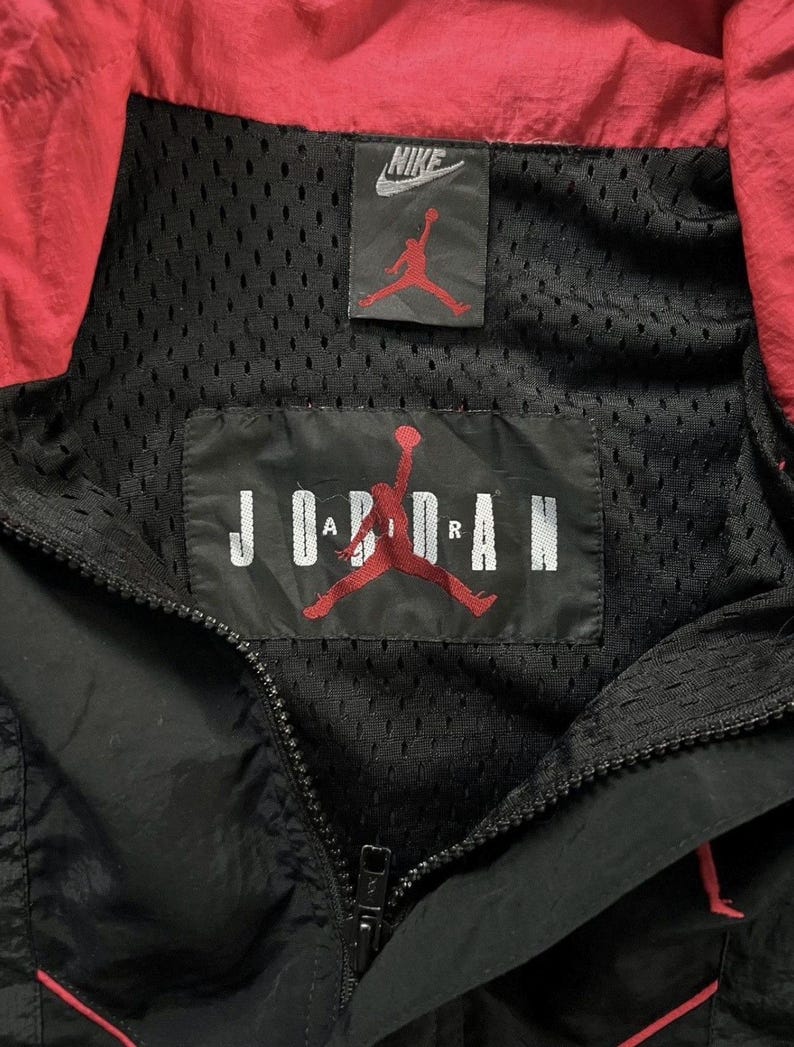 May include: Black and red Nike Jordan jacket. The jacket has a black mesh lining, a red collar, and a zip closure. The Nike logo and the Jordan logo are visible on the jacket.